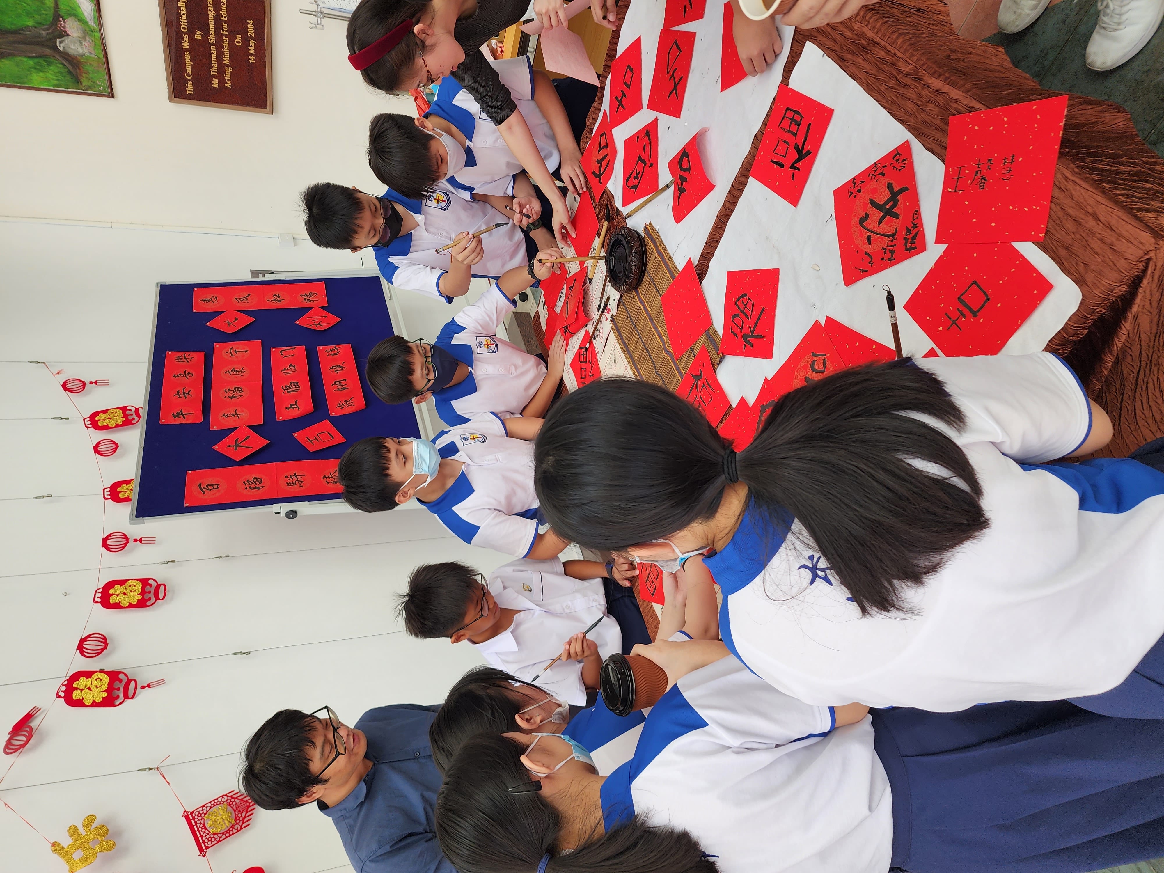 Students in school uniforms practice Chinese calligraphy on red paper around a table decorated with Lunar New Year ornaments.